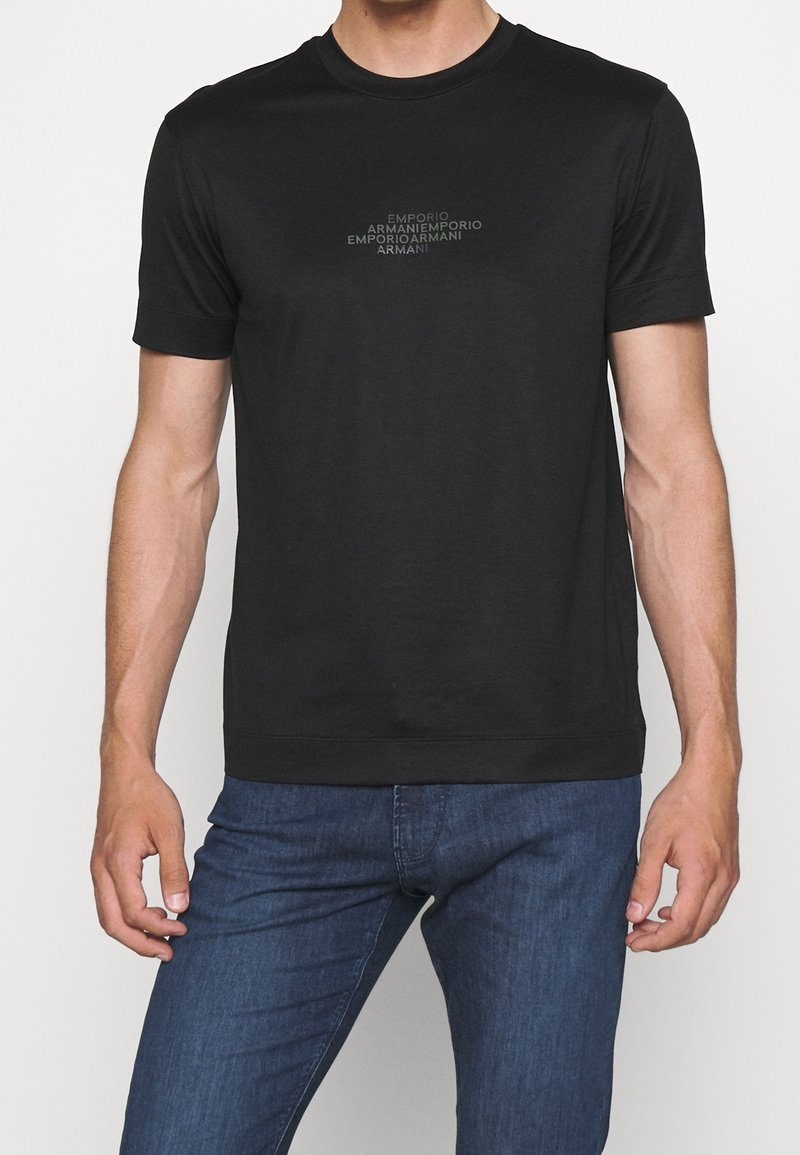 Svart t-shirt med rund hals i bomull, med en centrerad logotryck som har upprepade varunamn i en subtil grå nyans. Korta ärmar.