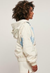 Lichte, korte jas in crème en lichtblauw, met een trekkoordcapuchon, elastische manchetten en een kleurgeblokt ontwerp. Gladde textuur.