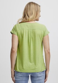 Camicia verde a maniche corte con finitura testurizzata, caratterizzata da un collo arrotondato e un dettaglio di piega sul retro, abbinata a jeans azzurri chiari.