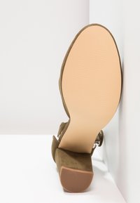 Chaussure femme en daim marron avec talon bloc et bride à la cheville, semelle face à l'appareil photo sur fond blanc.