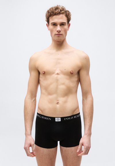 Νεαρός άνδρας με σγουρά μαλλιά, φορώντας μαύρα boxer briefs Polo Ralph Lauren, στέκεται μπροστά σε απλό φωτεινό φόντο, στραμμένος προς τα εμπρός.