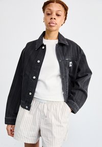 Giacca di denim nera con righe verticali, grande colletto e bottoni argentati. T-shirt bianca sotto, abbinata a pantaloni corti chiari a righe.