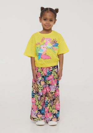 T-shirt giallo brillante con una grafica di due pappagalli, abbinato a pantaloni larghi con un motivo floreale colorato su sfondo nero.