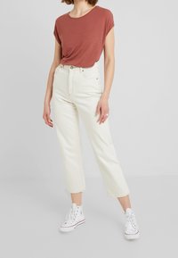 Kobieta w brązowej bluzce z krótkim rękawem, wysokich beżowych dżinsach i białych trampkach stojąca na jednolitym tle.