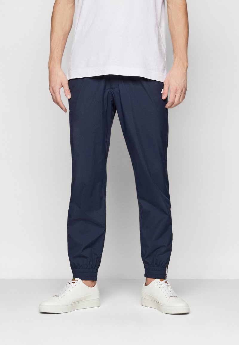 Homme portant un pantalon de jogging bleu marine et des baskets blanches, debout, les bras détendus le long du corps devant un fond uni clair.