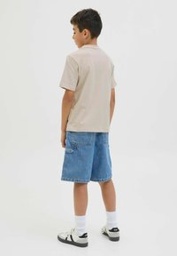 Šviesiai ruda trumpomis rankovėmis marškinėliai derinami su mėlynomis džinsinėmis šortais. Šortai turi dvi kišenes gale ir siekia šiek tiek virš kelių. Modelis dėvi baltas kojines ir juodus sportinius batus.