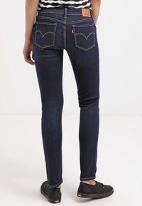 Jeans oscuros de corte Slim-fit con una cintura de altura media, dos bolsillos traseros y detalles de costura distintivos. Llevados con zapatos negros.