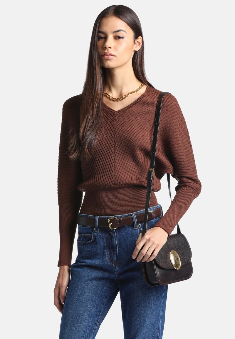 Femme portant un pull côtelé marron, un jean bleu, un collier chaîne en or et portant un sac en cuir noir porté en bandoulière avec une fermeture dorée.
