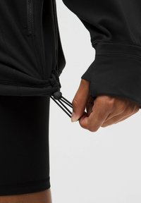 Čierna športová bunda so zipsom, nastaviteľnou šnúrkou na leme a otvormi na palce na rukávoch. Detailný záber ruky ťahajúcej za šnúrku.