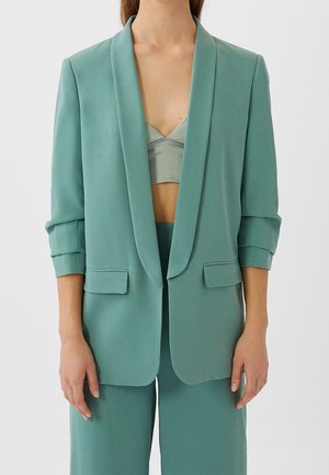 Vrouw die een teal blazer draagt met opgerolde mouwen, over een bijpassende cropped top en wijde broek, staand tegen een effen witte achtergrond.