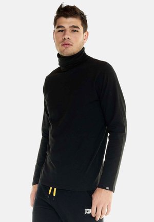 Jeune homme portant un col roulé noir à manches longues et un pantalon noir avec des cordons jaunes, debout devant un fond clair uni.