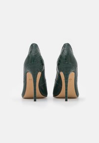 Une paire de chaussures à talons hauts vertes foncées avec une texture crocodile, vue de l'arrière sur un fond blanc.