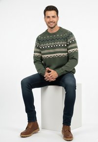 Suéter verde de punto con patrones geométricos en crema y azul marino, combinado con jeans oscuros y botas marrones con cordones sobre un pedestal blanco.