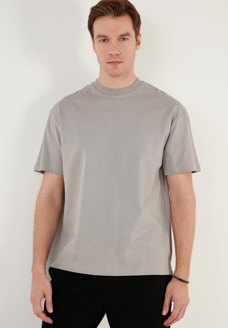 Grå t-shirt med korte ærmer i et blødt stof, med en rund halsudskæring og afslappet pasform, båret med mørke bukser og hænderne ned langs siden.