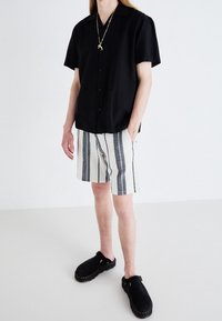 Person som bär en svart kortärmad skjorta med knäppning, randiga vita och svarta shorts, svarta slip-on skor och ett guldhänge.