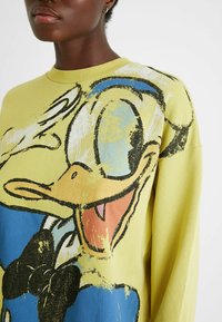 Vaaleankeltainen collegepaita, jossa on sarjakuvatyylinen ankka-kuvio. Kuosissa on sinisiä ja mustia yksityiskohtia sekä teksturoitu pinta.