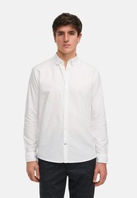 Chemise blanche à manches longues avec un col, texture lisse et coupe décontractée. Présente une patte de boutonnage et des boutons sur le devant.