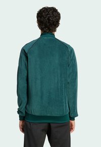 Uomo con capelli ricci che indossa una giacca a maniche lunghe turchese e pantaloni neri, di spalle su uno sfondo chiaro e semplice.