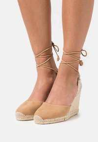 Dei tacchi a wedge in suede beige con suola in juta intrecciata e lacci decorativi alla caviglia. Presentano una punta arrotondata e una superficie texture.