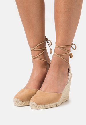 Dei tacchi a wedge in suede beige con suola in juta intrecciata e lacci decorativi alla caviglia. Presentano una punta arrotondata e una superficie texture.