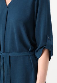 Blouse bleu sarcelle avec un col en V, manches retroussées ornées de boutons, et ceinture nouée à la taille. Tissu doux et texturé.