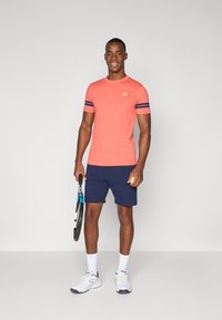 Korallfarget t-skjorte med kort erme og marineblå striper, kombinert med marineblå shorts. Holder en tennisketcher og en gul ball, iført hvite joggesko.