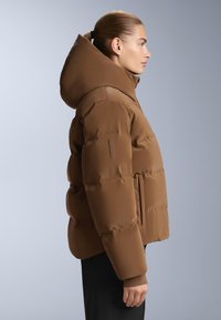 Smeđa puffer jakna s velikim kapuljačom, sadrži kvadratni uzorak, elastične manžete i bočnu džep, prikazana sa strane.