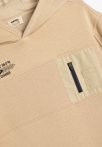 Sudadera beige con capucha, con una textura lisa, bolsillo frontal con cremallera negra y diseño impreso encima del bolsillo.