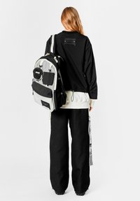 Mochila en blanco y negro con múltiples bolsillos y cremalleras; tela texturizada; combinada con una camiseta de manga larga negra y pantalones negros holgados.