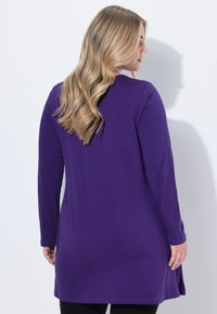 Fioletowa tunika z długim rękawem o luźnym kroju, gładkiej fakturze i bocznych rozcięciach; noszona na czarnych legginsach, na modelce odwróconej tyłem.