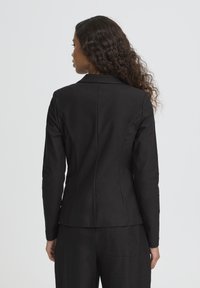 Blazer noir cintré avec une coupe ajustée, des revers crantés et une texture lisse. Il présente une couture centrale au dos et des manches longues.