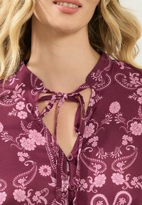 Blusa bordeaux con design floreale, colletto a fiocco e chiusura con bottoni. Presenta fiori rosa e motivi paisley su tutta la superficie.