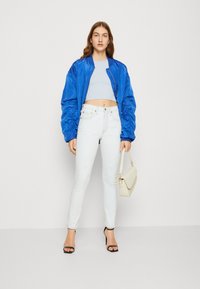 Kék selyem bomber dzseki, világoskék cropped top, koptatott fehér farmert, fekete pántos magassarkú cipőt, és egy krém színű kézitáskát visel. Egyszerű háttér.
