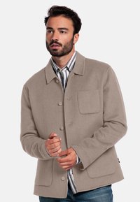 Veste en mélange de laine beige avec un devant à boutons, une poche poitrine unique et des manches droite. Portée sur une chemise rayée.