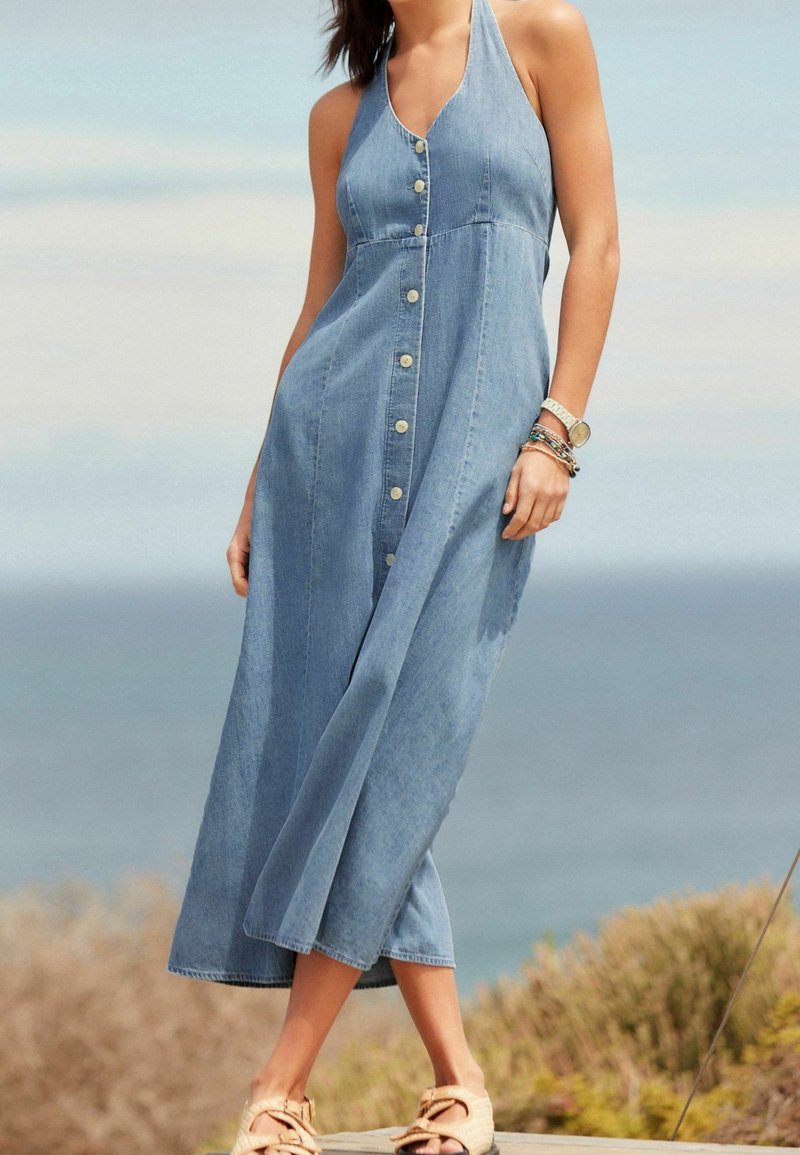 Robe maxi en denim avec décolleté halter, fermeture à boutons sur le devant et coupe décontractée. Couleur bleu clair, texture douce et design fluide.