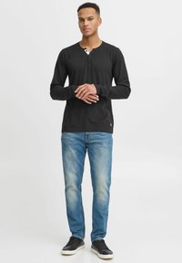Fekete, hosszúujjú Henley ing, gombos nyakkivágással, világoskék farmernadrággal és fekete sportcipővel, semleges háttér előtt.