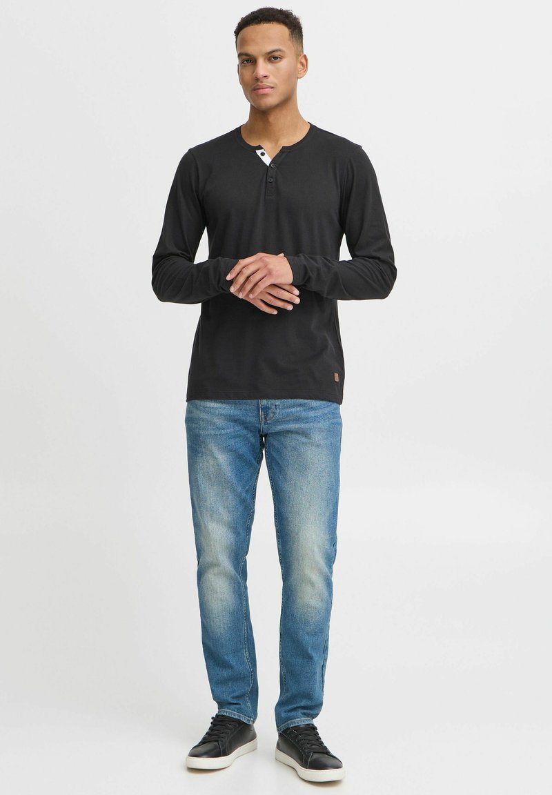 Schwarzes Langarm-Henley-Shirt mit knöpfbarem Ausschnitt, kombiniert mit hellblauen Jeans und schwarzen Turnschuhen vor einem neutralen Hintergrund.
