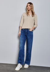 Beige Langarmoberteil mit durchgehender Knopfleiste, kombiniert mit blauen weit geschnittenen Jeans und weißen Sneakers vor einem grauen Hintergrund.