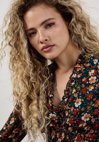 Bluse mit Blumenmuster in Dunkelgrün, rüschigem Halsausschnitt und langen Ärmeln. Beinhaltet kleine Blumen in Rot, Weiß und Orange auf einem transparenten Stoff.