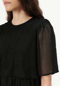 Crna haljina s kratkim, prozirnim rukavima, okruglim izrezom i slojevitim, lepršavim dizajnom. Tkanina ima mekan i lagan teksturu.