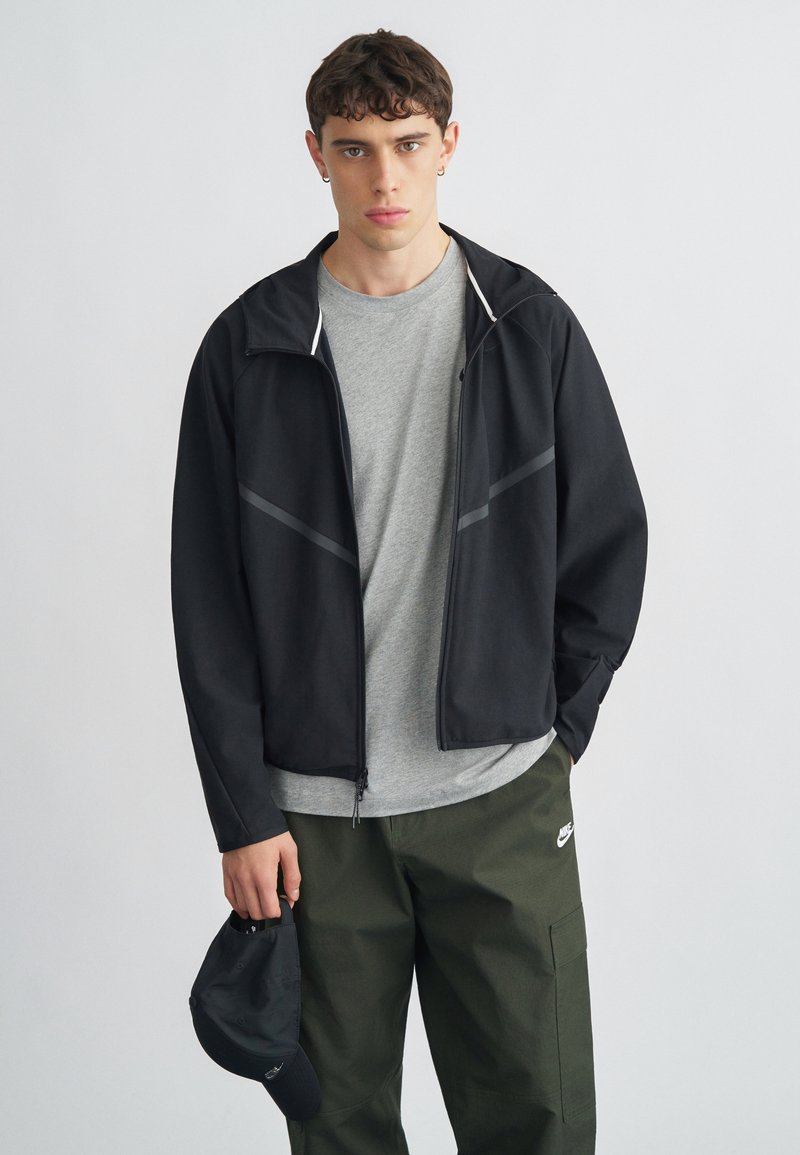 Jaqueta preta leve com detalhes cinza, fecho frontal com zíper, combinada com uma t-shirt cinza e calças cargo verde-oliva, segurando um boné preto.