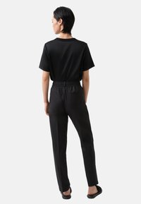 Zwarte shirt met korte mouwen, gecombineerd met zwarte elastische broek; gladde textuur, relaxed fit, subtiele achterzakdetail en platte sandalen.