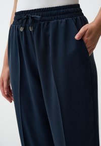 Marineblauwe, elastische broek met verstelbare elastische band en zijzakken. De stof heeft een gladde textuur met een subtiele glans.