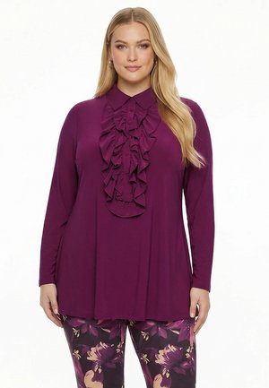 Kvinde iført en langærmet lilla bluse med flæser ned foran og bukser med blomsterprint i lilla og beige toner.