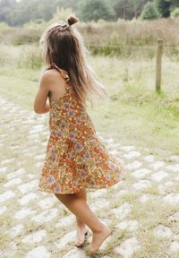 Oranje katoenen jurk met bloemmotieven, gerimpelde schouderbanden en een gelaagde rok. Blotevoets kind dat rent op een stenen pad.