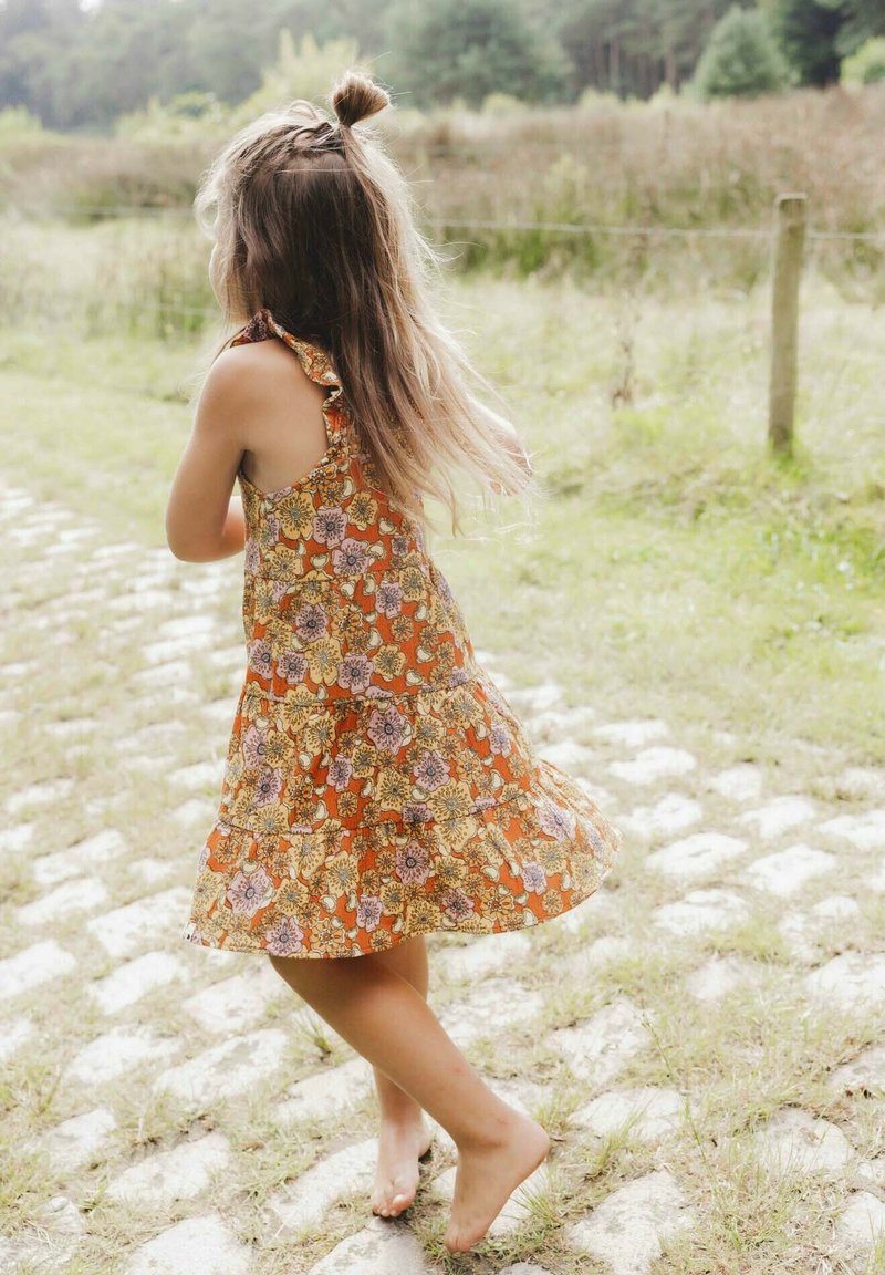 Oranje katoenen jurk met bloemmotieven, gerimpelde schouderbanden en een gelaagde rok. Blotevoets kind dat rent op een stenen pad.