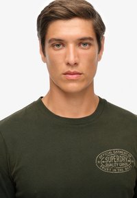 Camiseta de algodón verde oliva con cuello redondo y mangas largas, que presenta un logo impreso en un tono beige claro en el pecho. Textura suave.