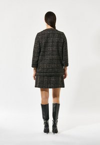 Schwarz-braues, strukturiertes kariertes Kleid mit Dreiviertelärmel und geradem Saum, kombiniert mit kniehohen schwarzen Stiefeletten mit Absatz.