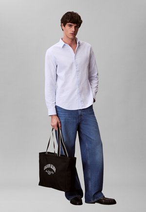 Muškarac u svijetloplavoj košulji s kopčanjem i širokim trapericama, drži crnu Calvin Klein torbu s bijelim tekstom.