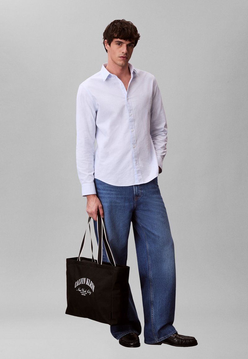 Άνδρας που φοράει ανοιχτό μπλε πουκάμισο με κουμπιά και τζιν με φαρδύ παντελόνι, κρατώντας μια μαύρη τσάντα tote Calvin Klein με λευκό κείμενο.